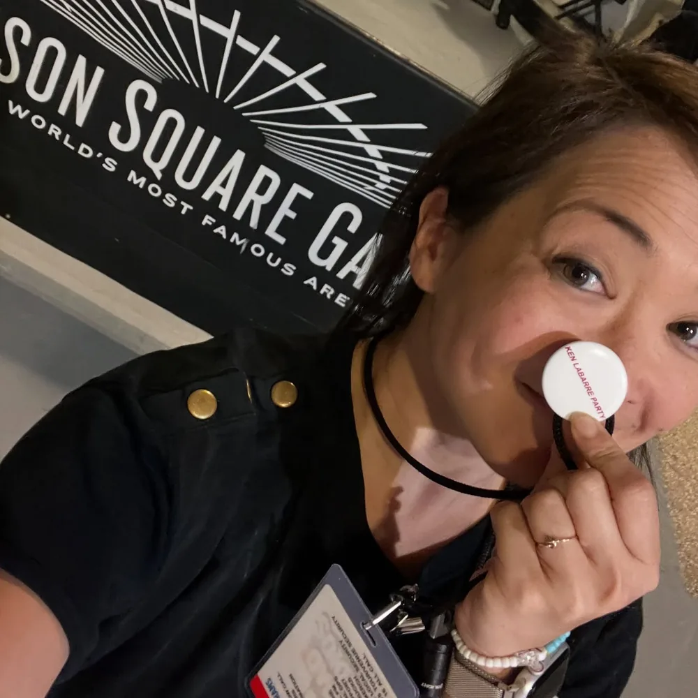 Selena Moshell backstage at an arena with her crew pass, holding a round button to her cheek beside an arena banner