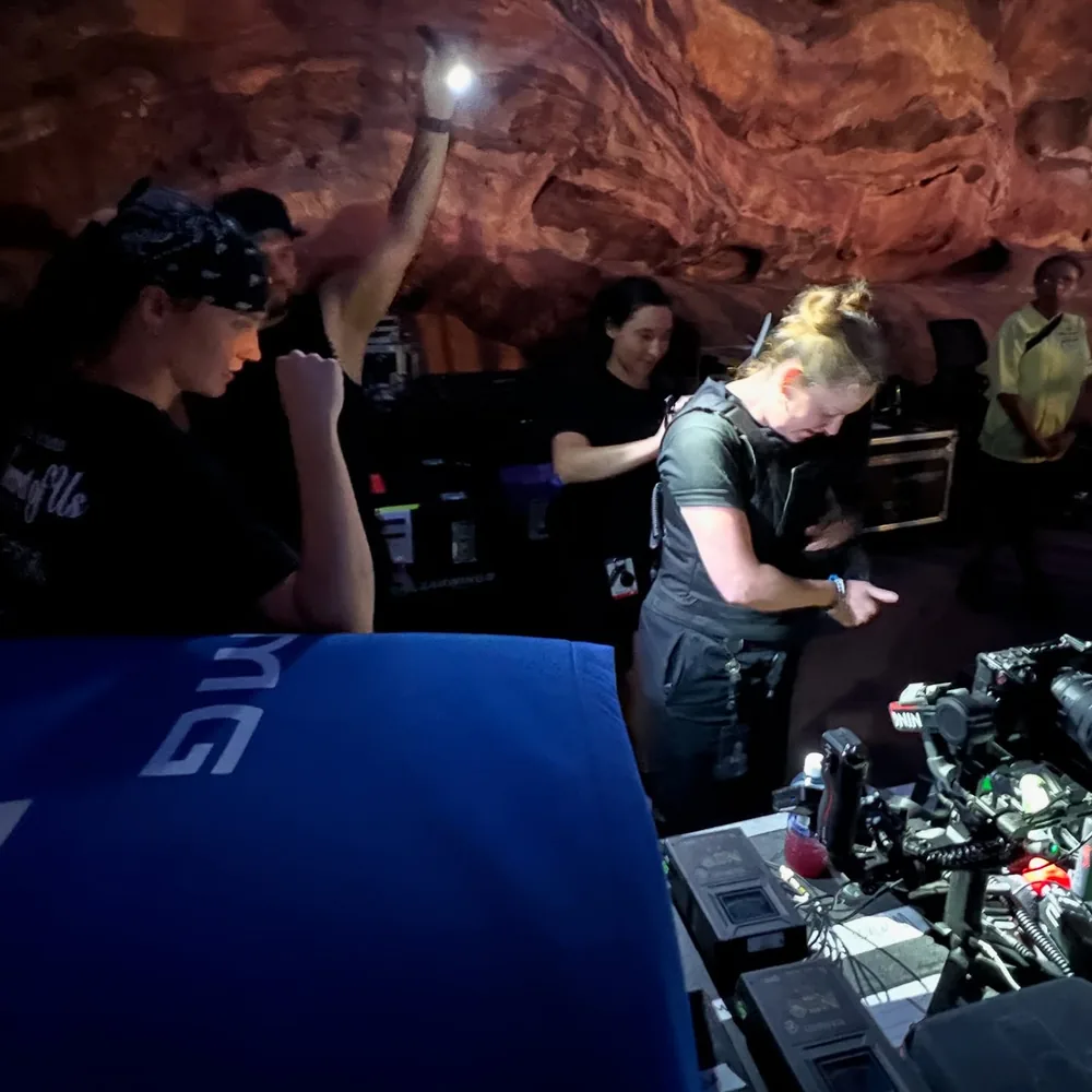 Show crew prepping cameras and a gimbal at a lit desk under a towering red rock wall at night