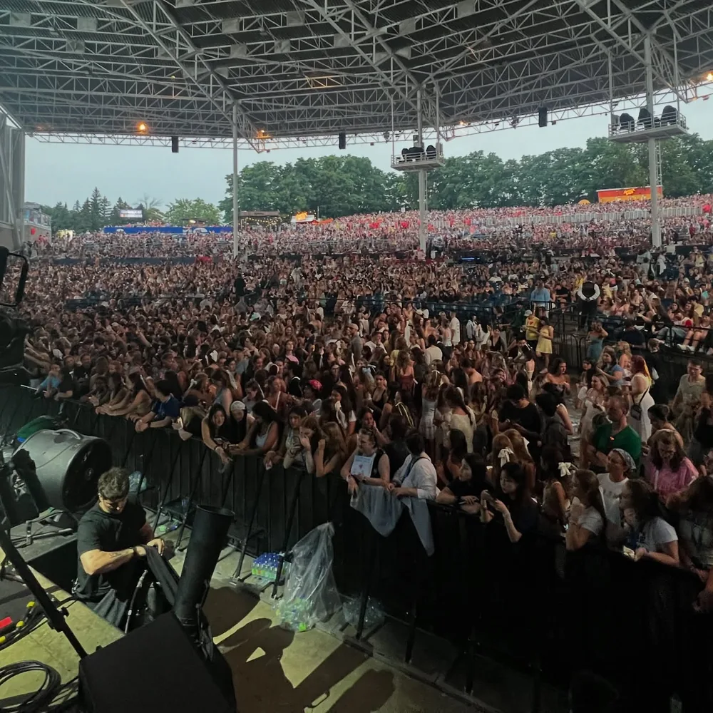 Packed amphitheater crowd seen from the stage under a steel roof, with a crew member working at the barricade